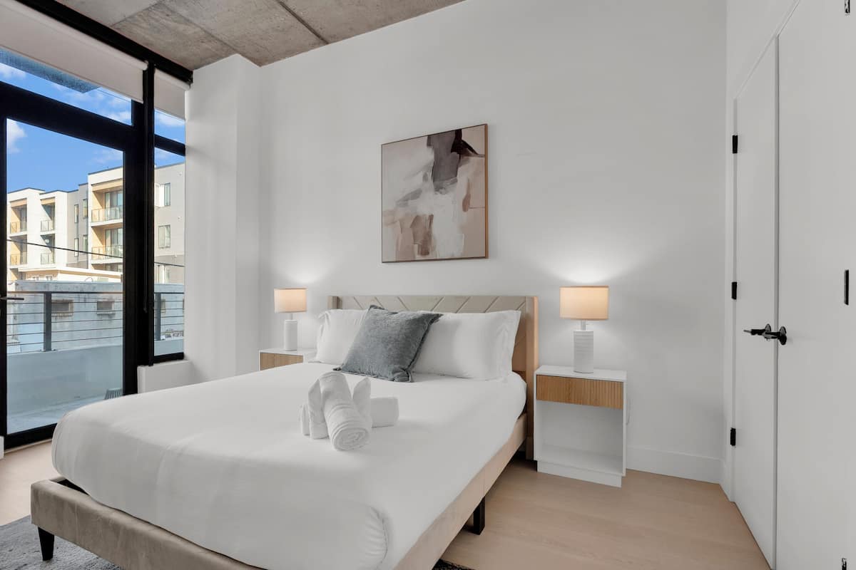 A modern bedroom with a large bed, two bedside tables, a nightstand, a painting, and a view through a large window.