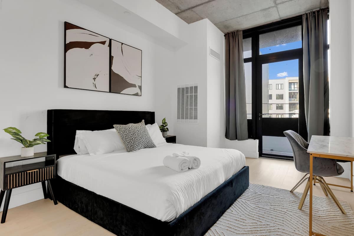 A modern bedroom with a large bed, a desk, and a view of buildings through a window.