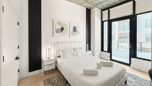 A modern bedroom with a large bed, a nightstand, a wall-mounted light, and a view of the city through large windows. - Apartmint at N 2nd St Philadelphia (Philadelphia)