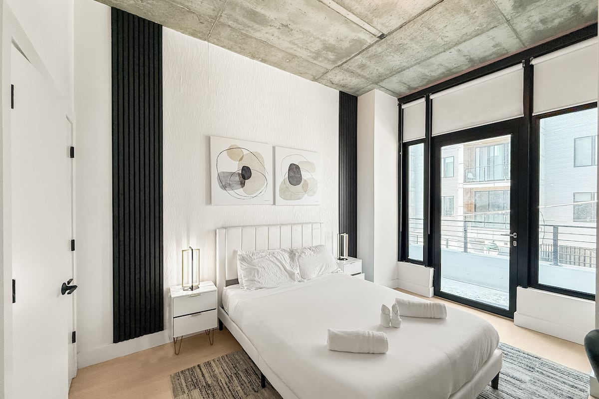 A modern bedroom with a large bed, a nightstand, a wall-mounted light, and a view of the city through large windows.