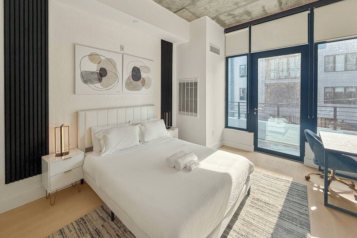 A modern bedroom with a large bed, a nightstand, a desk, and a view of the city through large windows.