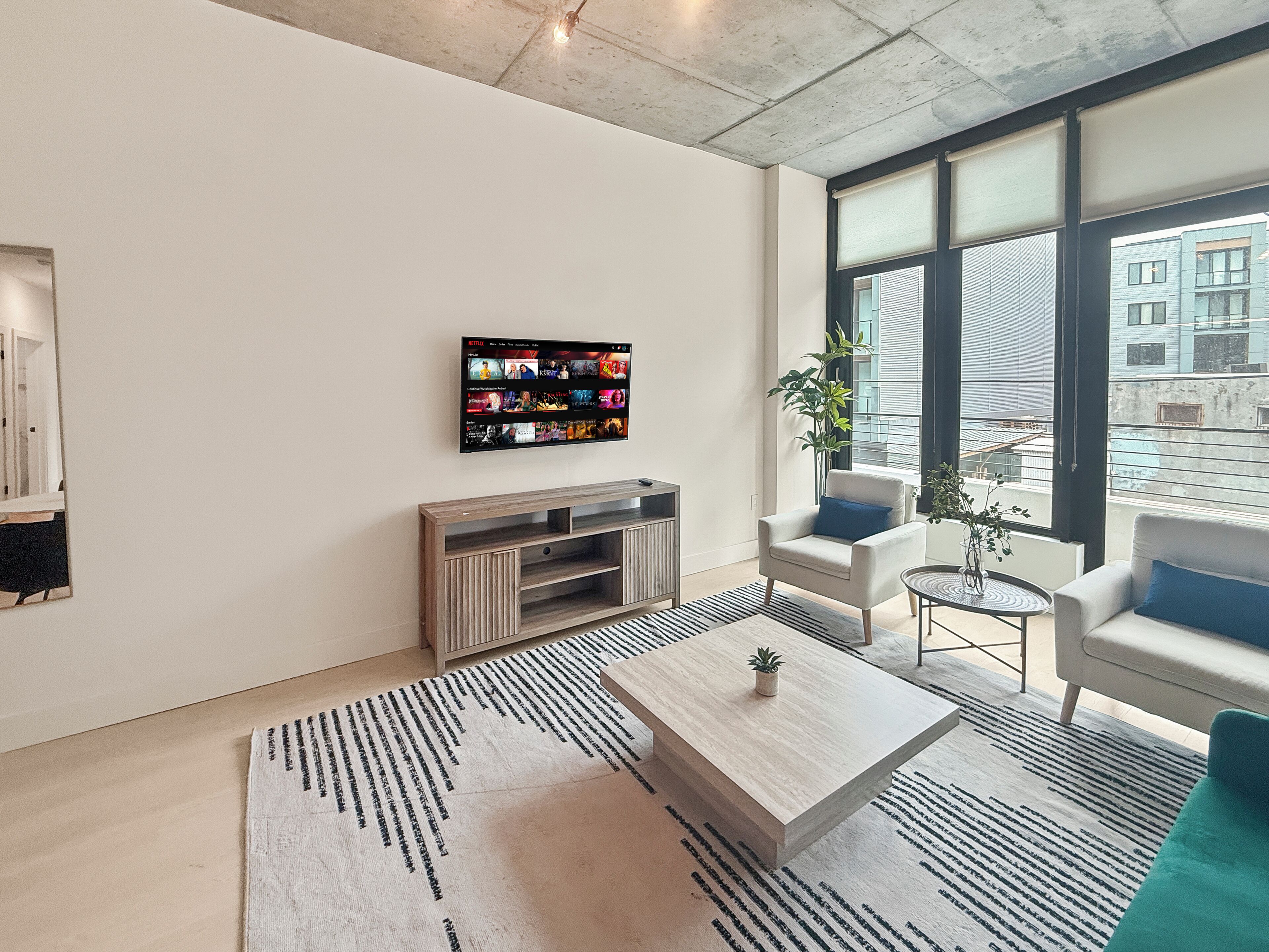 A modern living room with a flat-screen TV, a wooden entertainment unit, a coffee table, a sofa, and a large window with a view of buildings.