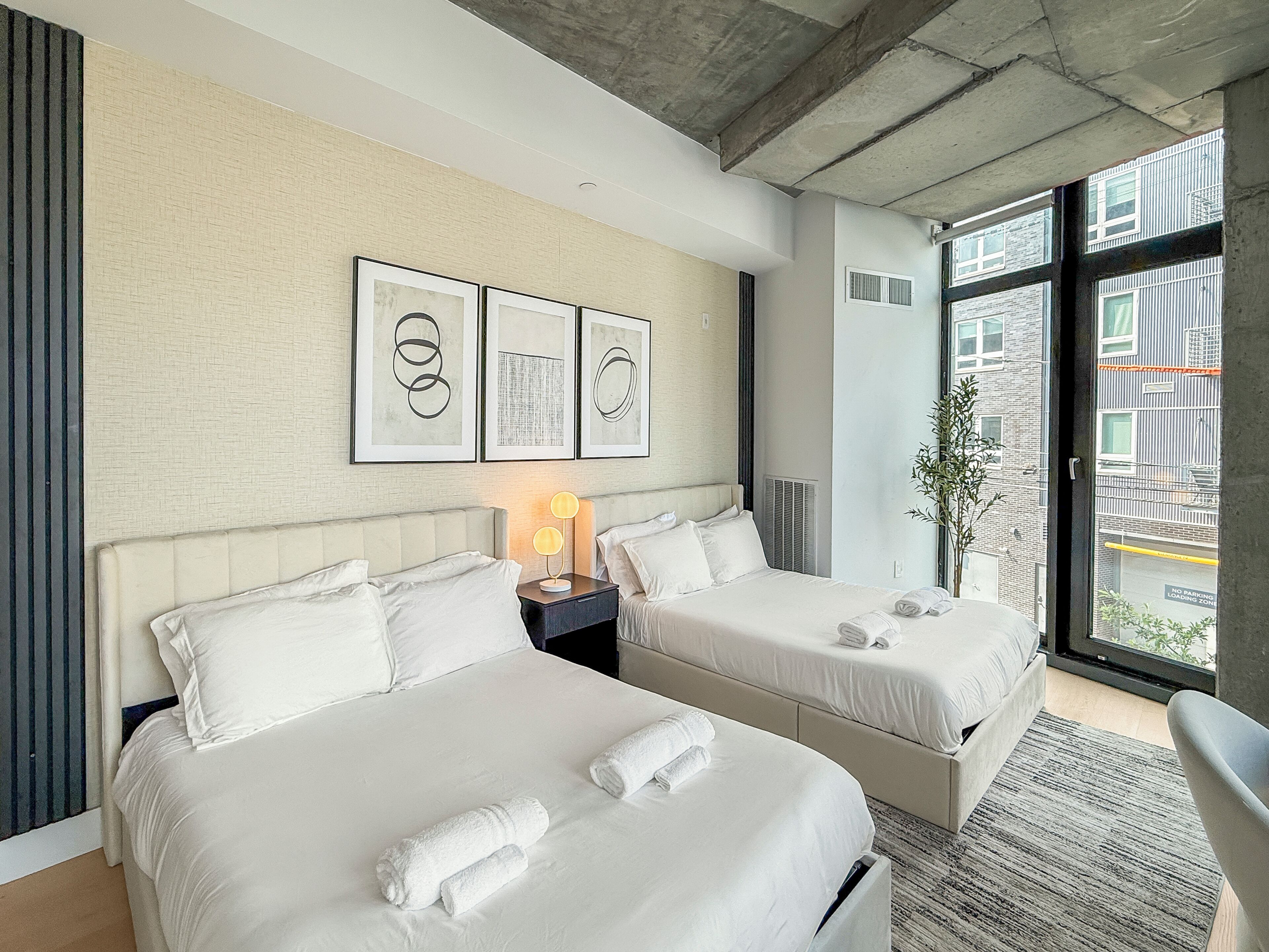 A hotel room with two beds, a nightstand with a lamp, framed artwork on the wall, a potted plant, and a view of the outside building.