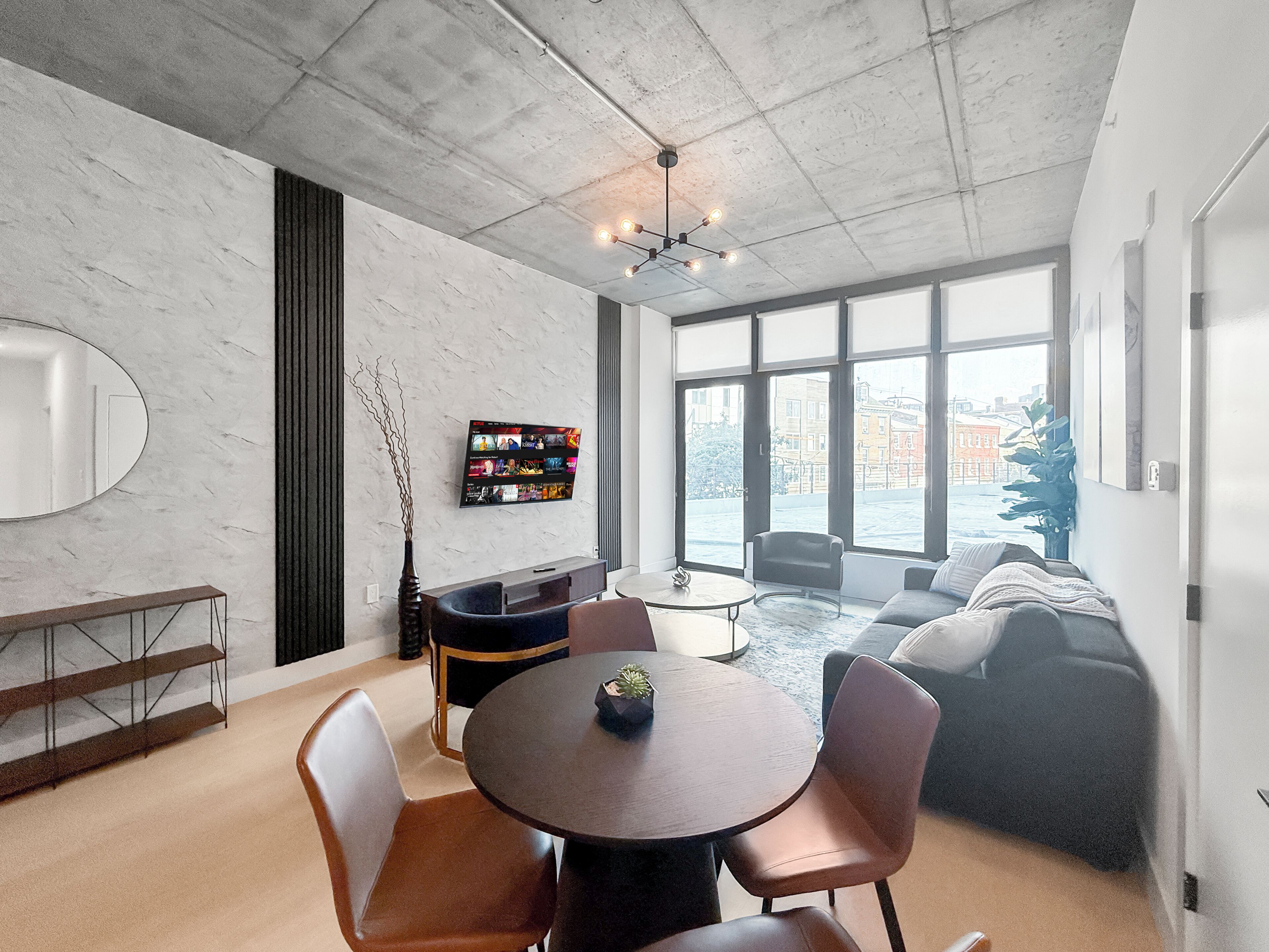 A modern living room with a round table, chairs, a sofa, a TV mounted on the wall, and large windows.