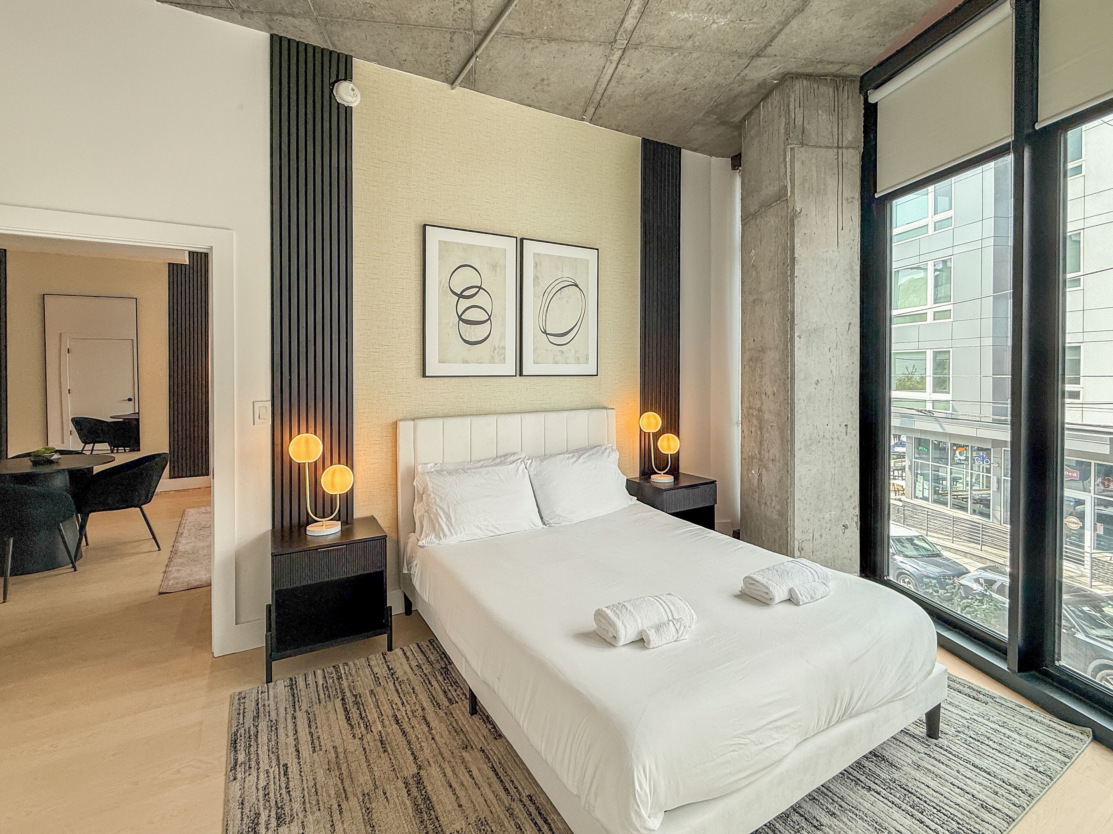 A modern hotel room with a large bed, two bedside lamps, a view of the city, and a dining area.