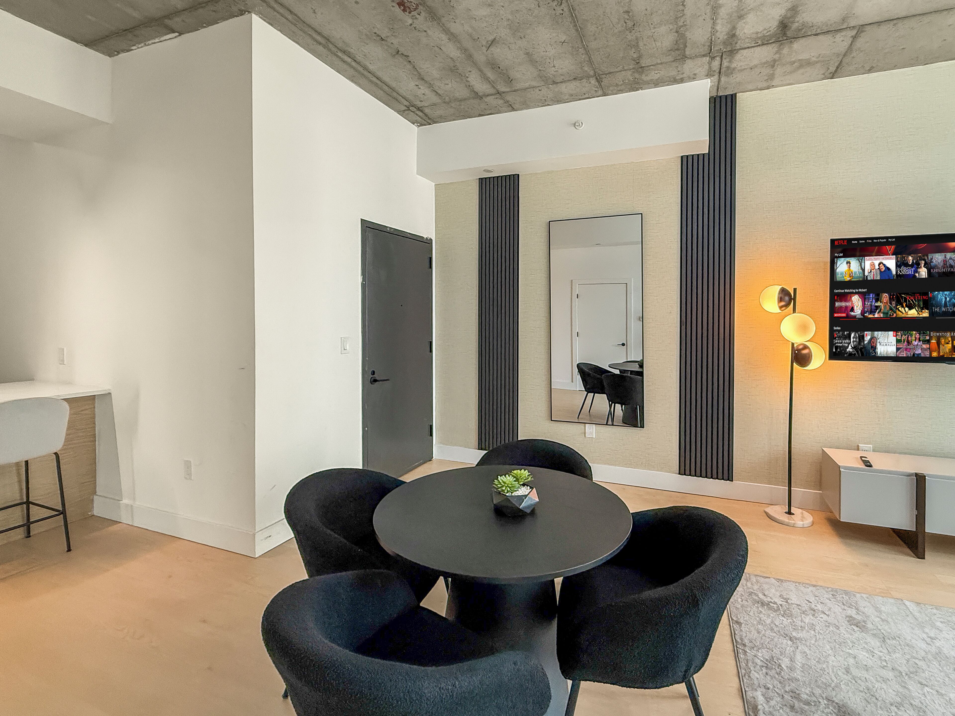 A modern living room with a round table, black chairs, a small plant, a large mirror, a TV wall mounted on the wall, and a minimalist design.
