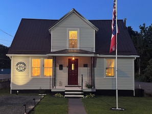A two-story house with a red door, a flagpole with a flag, and a small porch with steps.