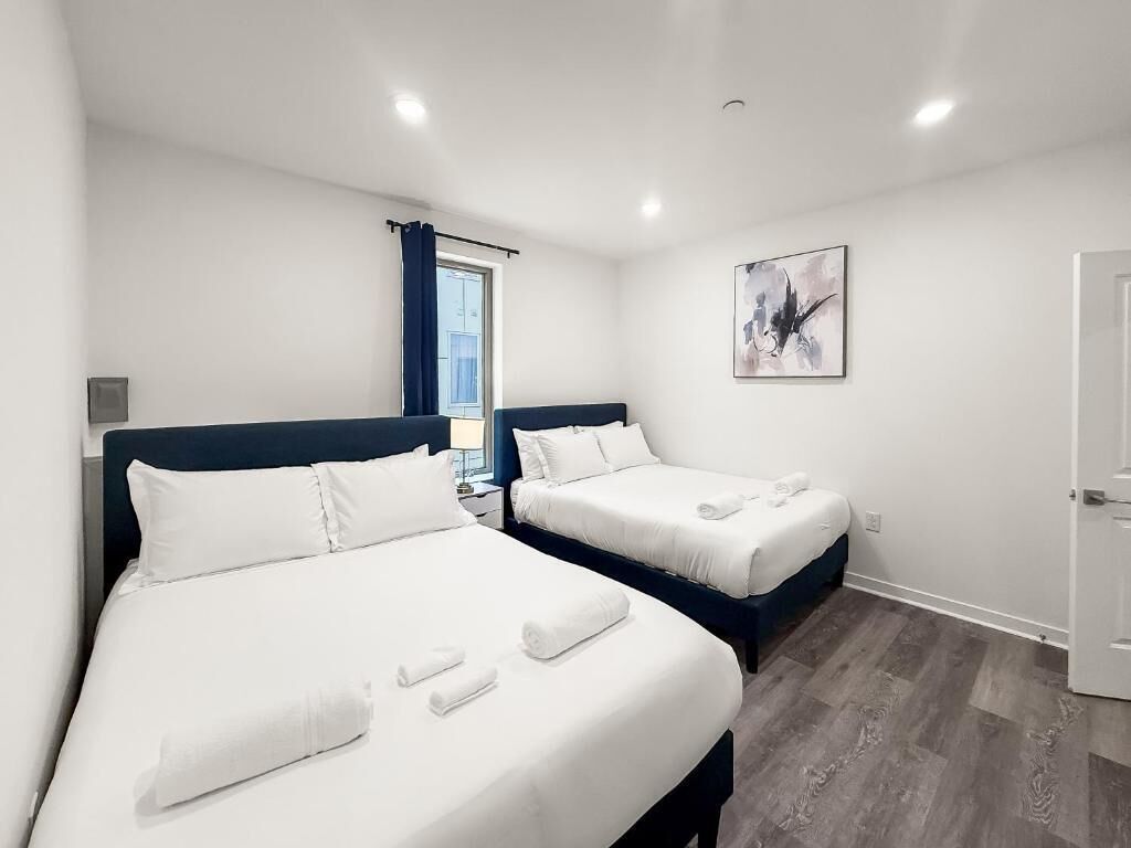 Camera d'albergo con due letti, una finestra con tende blu, un quadro appeso al muro e una porta.