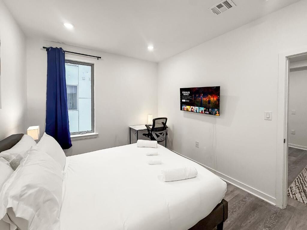 A hotel room with a bed, a desk, a chair, a TV on the wall, and a window with blue curtains.