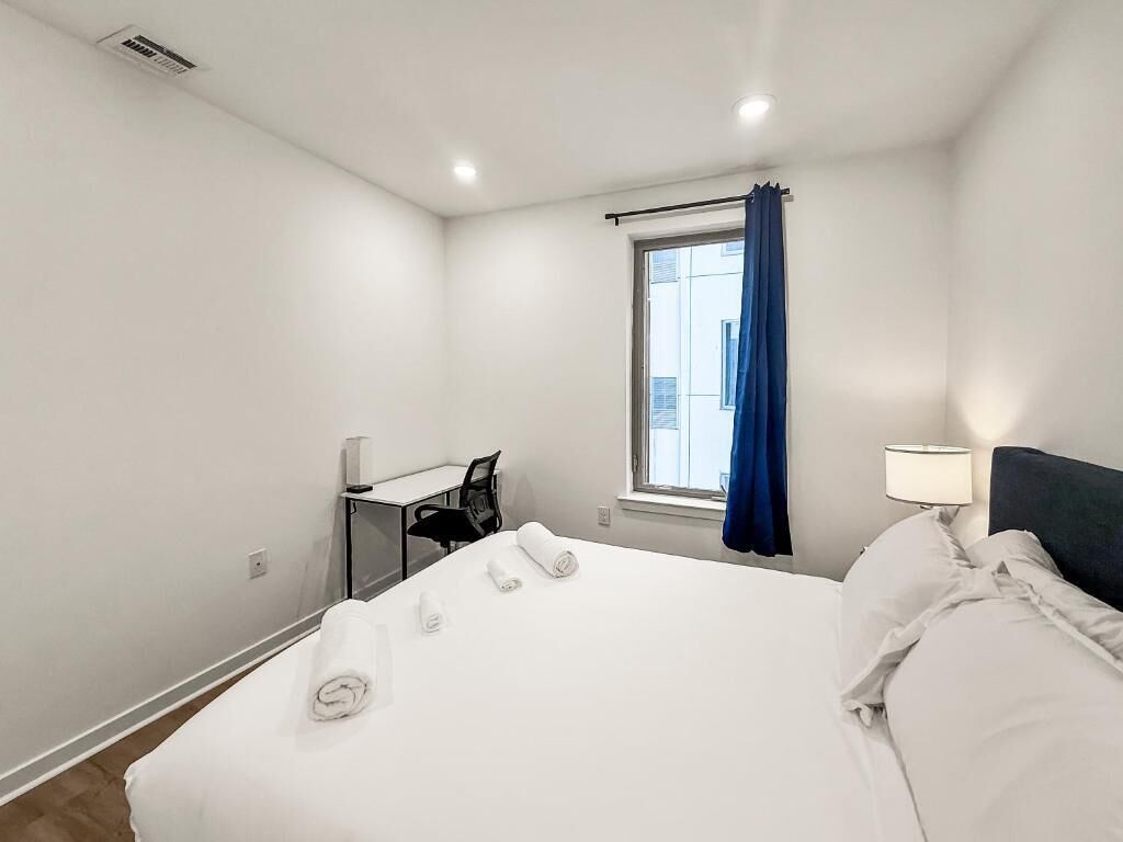 A hotel room with a bed, a desk, a chair, a window with curtains, and a nightstand with a lamp.