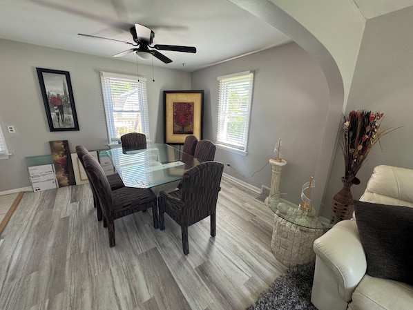 A dining area with a glass table, dark chairs, a ceiling fan, framed artwork, and a vase with dried plants.