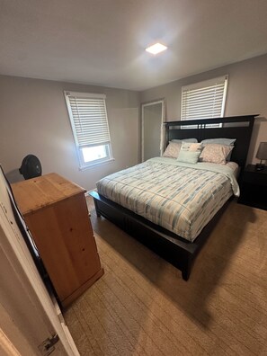 Ein Schlafzimmer mit Bett, Nachttisch, Kommode und zwei Fenstern mit Jalousien.