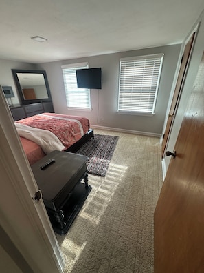 A bedroom with a bed, a dresser, a TV mounted on the wall, and two windows with blinds.
