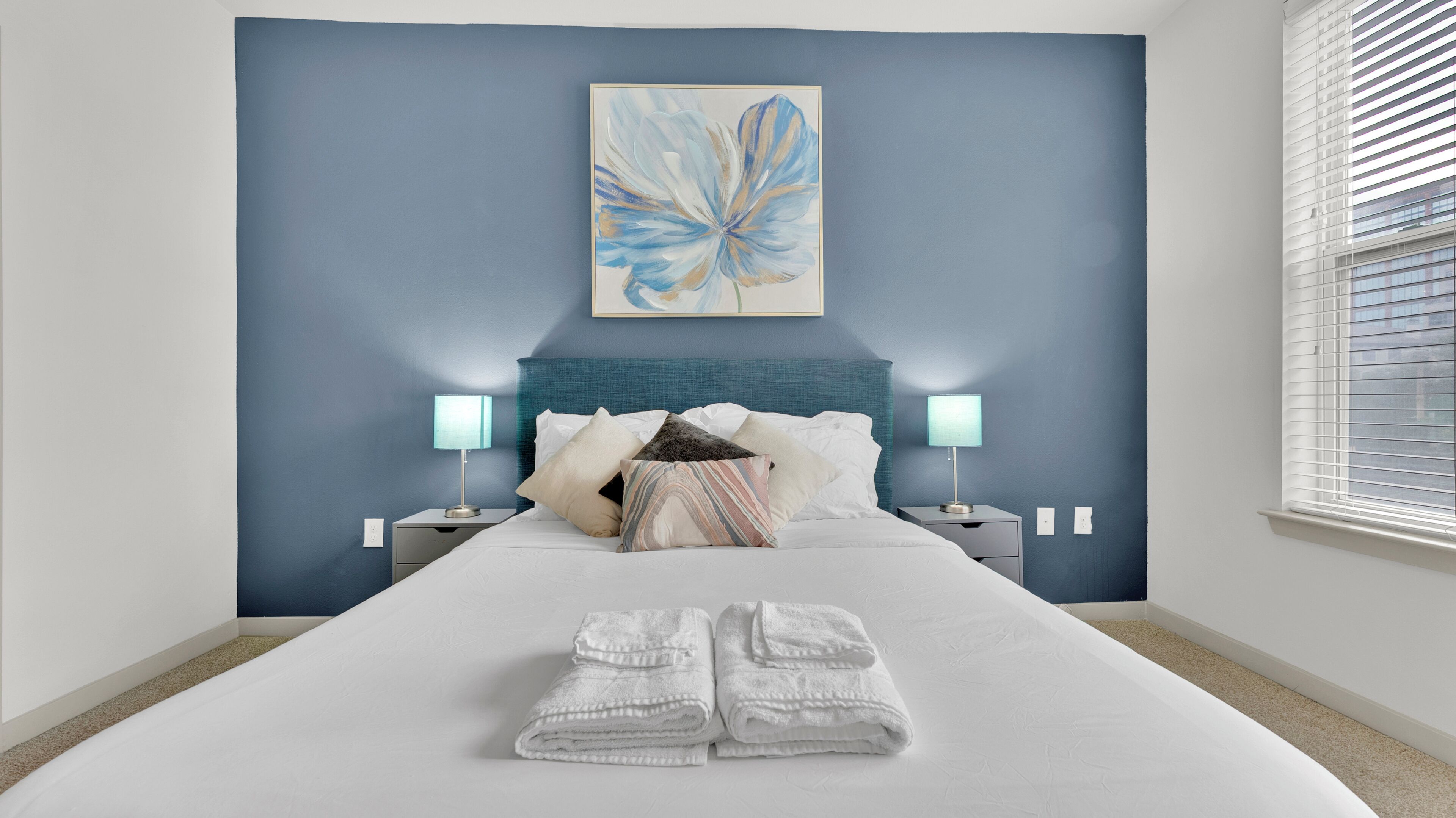 A hotel room with a bed, two bedside lamps, a blue wall, and a painting.