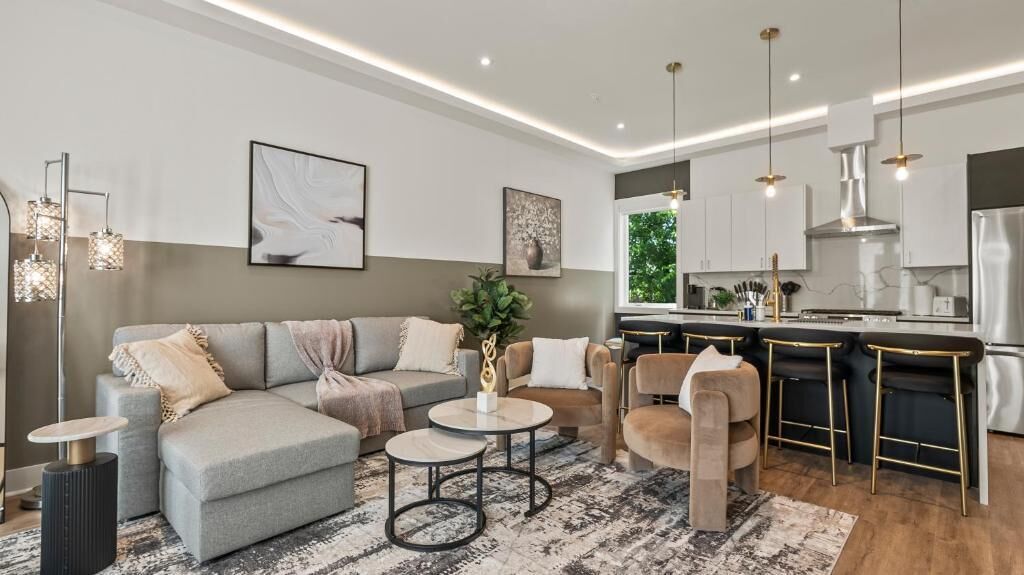 A modern living room with a sofa, coffee table, and chairs, a kitchen with a stove and refrigerator, and pendant lighting.