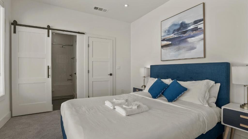 Une chambre à coucher comprenant un lit, une table de chevet, un tableau accroché au mur et une porte donnant sur une salle de bain.
