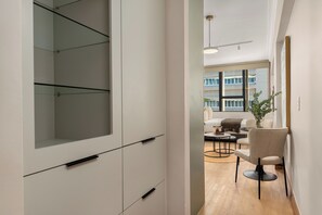 Ein modernes Interieur mit minimalistischem Design, ausgestattet mit einem Einbauschrank mit mehreren Schubladen und einer Glasablage, einer Sitzecke mit Sofa und Sessel sowie einem großen Fenster mit Blick auf die Außenansicht des Gebäudes.