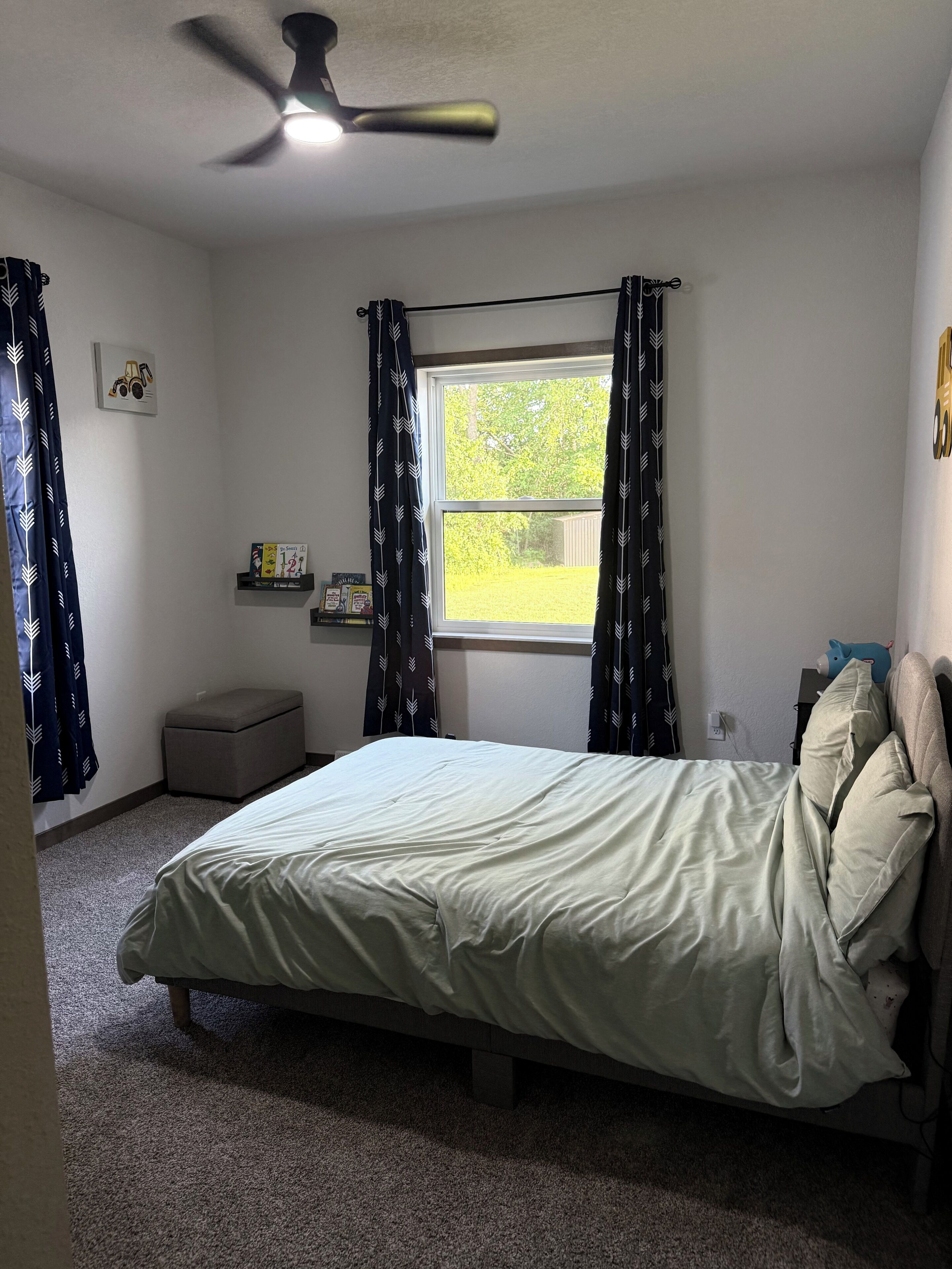 Un dormitorio con cama, ventilador de techo, ventana con cortinas, una pequeña banca y una estantería con libros.