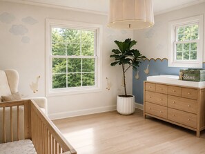 A room with a crib, a dresser, a potted plant, and a window with a view of trees.