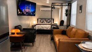 A compact hotel room with a bed, a sofa, a dining table, a TV mounted on the wall, and a kitchenette. - The Ember Suite l Designer Guest House (Kissimmee)