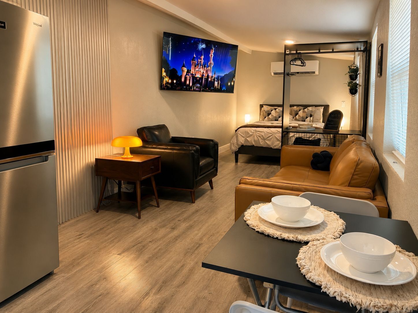 Una habitación de hotel compacta con kitchenette, una cama con cabecera, una zona de estar con sofá y sillas, un televisor montado en la pared y una ventana con persianas.