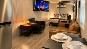 A compact hotel room with a kitchenette, a bed with a headboard, a seating area with a sofa and chairs, a television mounted on the wall, and a window with blinds. - The Ember Suite l Designer Guest House (Kissimmee)