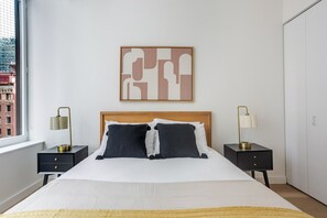 A bedroom with a bed, two bedside tables, a nightstand, a wall-mounted lamp, and a window with a view of buildings. - Blueground | Financial District, pool, nr Brooklyn Bridge (New York)