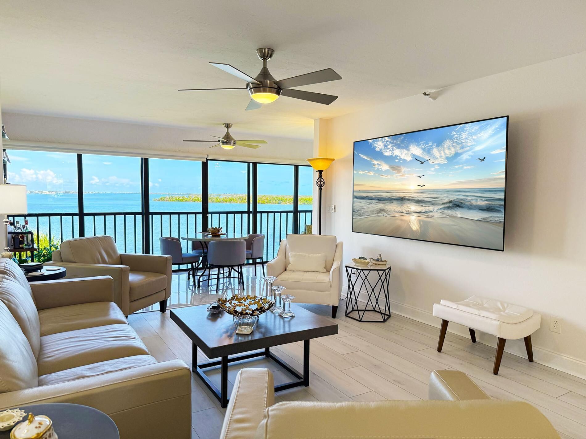Ein geräumiges Wohnzimmer mit großem Fenster mit Blick auf ein Gewässer, einem Deckenventilator, einem Couchtisch und einem Gemälde einer Strandlandschaft.