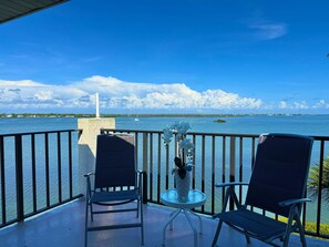 A balcony with two chairs and a small table, overlooking a body of water with a distant island and a sailboat. - Sandpiper Key Endless Bay Views & Pool (Englewood)