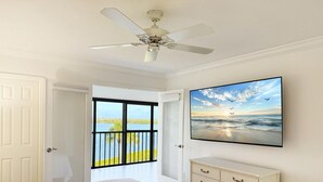 A bedroom with a bed, a dresser, a ceiling fan, a painting of a beach scene, and a door leading to a balcony with a view of water and sky. - Sandpiper Key Waterfront Views & Pool (Englewood)