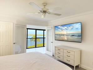 Ein Schlafzimmer mit Bett, Schminktisch, Deckenventilator, einem Strandbild und einer Tür zu einem Balkon mit Blick auf Wasser und Himmel.