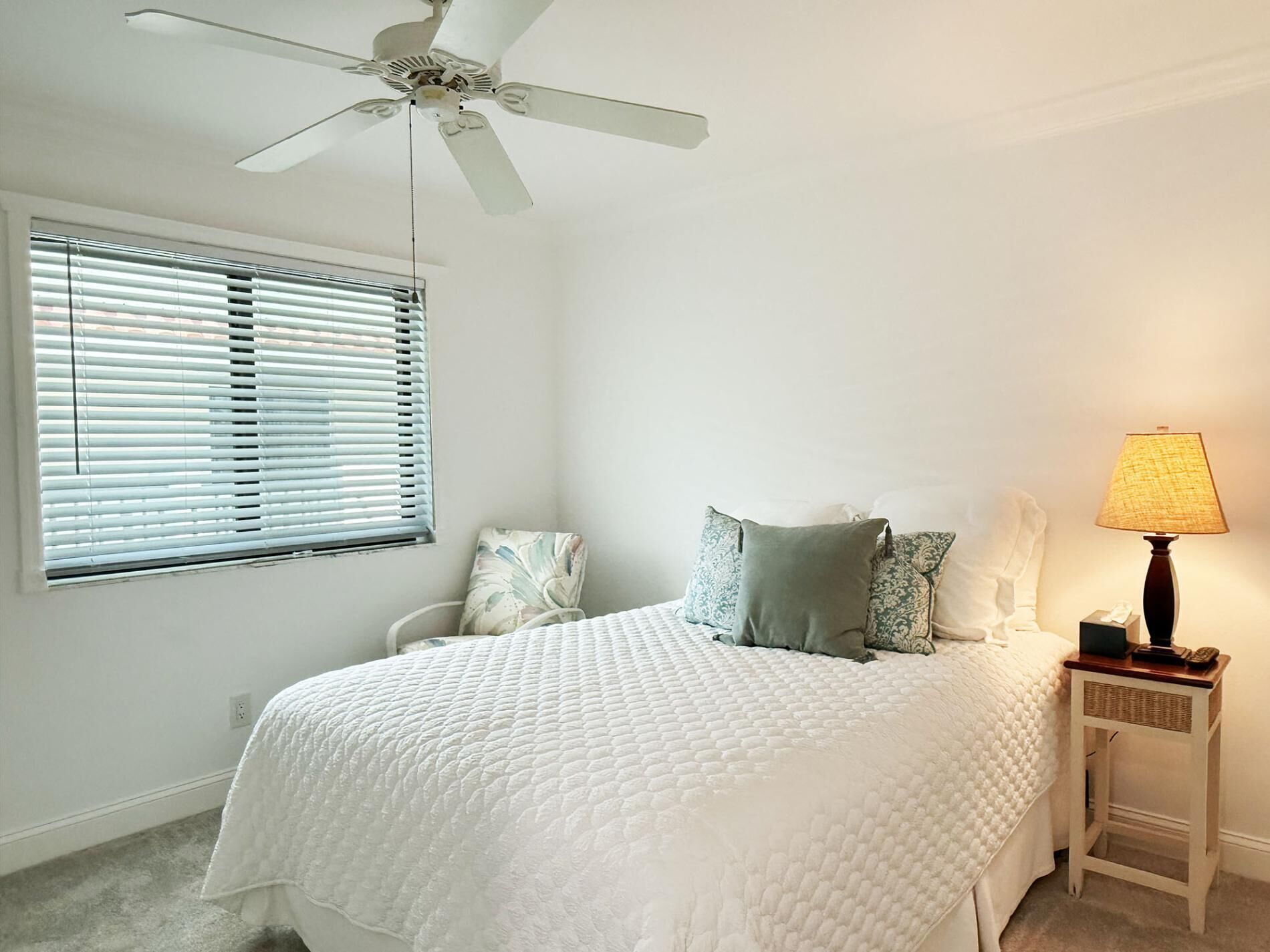 Une chambre à coucher comprenant un lit, une table de chevet, une lampe, un ventilateur de plafond et une fenêtre avec des stores.