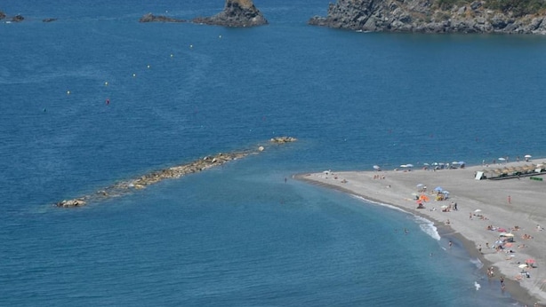 A coastal beach with clear blue waters, rocky outcrops, and a sandy shore with umbrellas.
