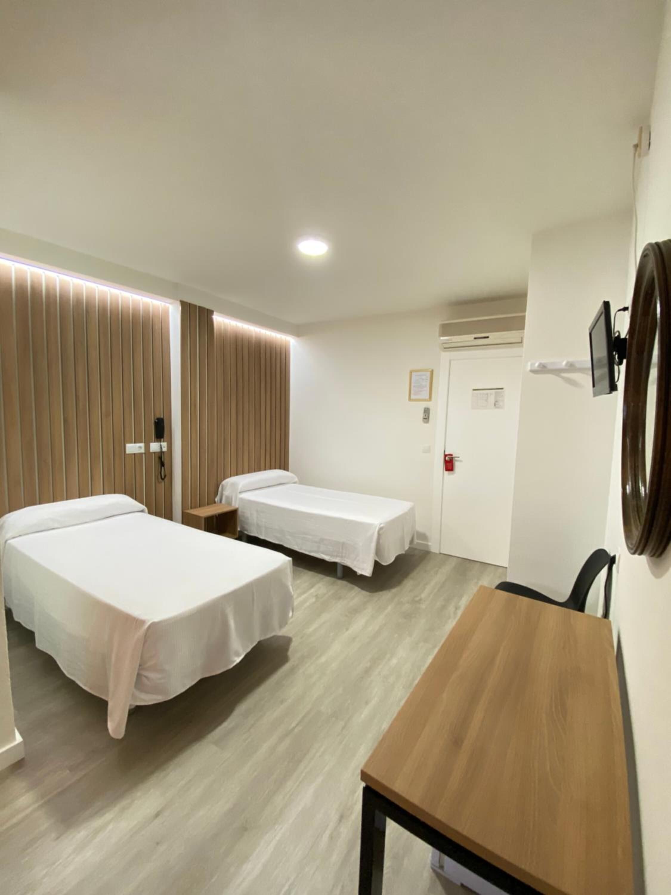 Ein Hotelzimmer mit zwei Betten, einem Holztisch, einem Schreibtisch, einem Spiegel und einer Tür mit Rauchmelder.