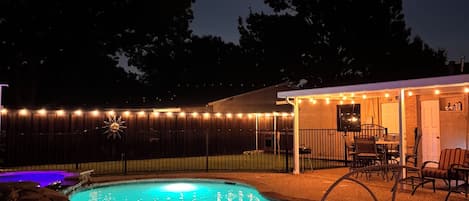 Uma piscina à noite com a lua no céu, um espaço coberto com espreguiçadeiras e um cercado com luzes de corda.
