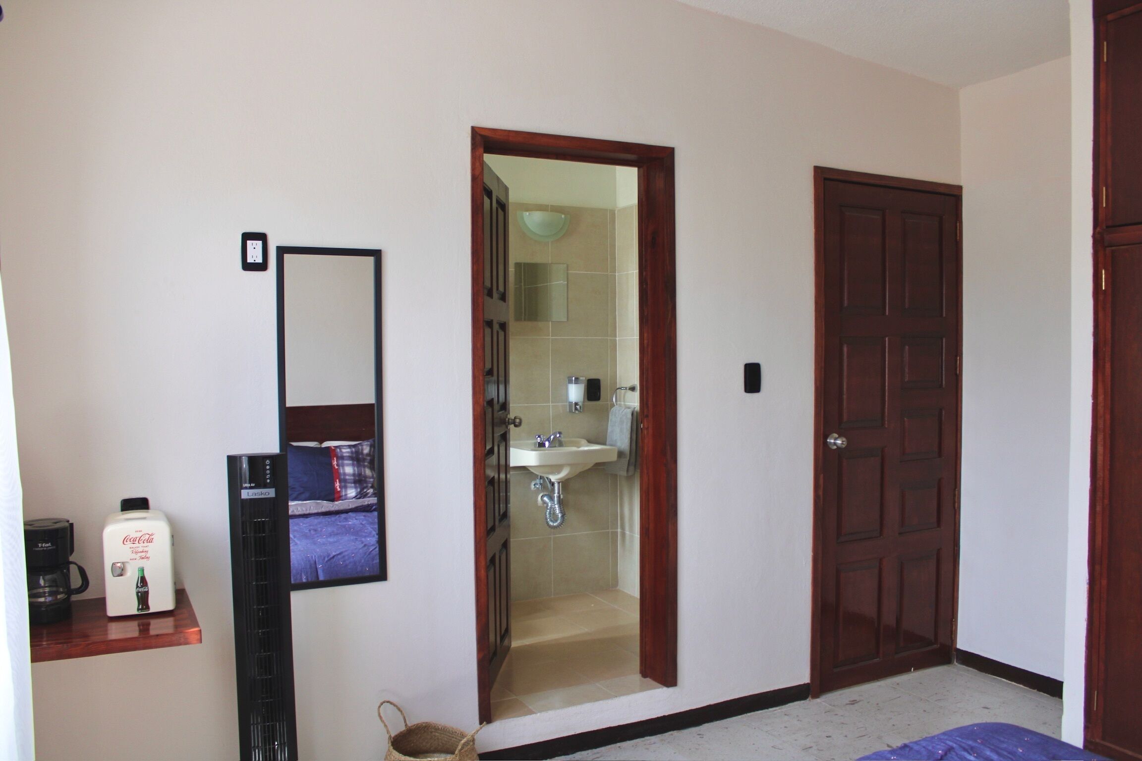 Habitación de hotel con cama, cafetera, espejo, puerta y baño visible a través de una puerta abierta.