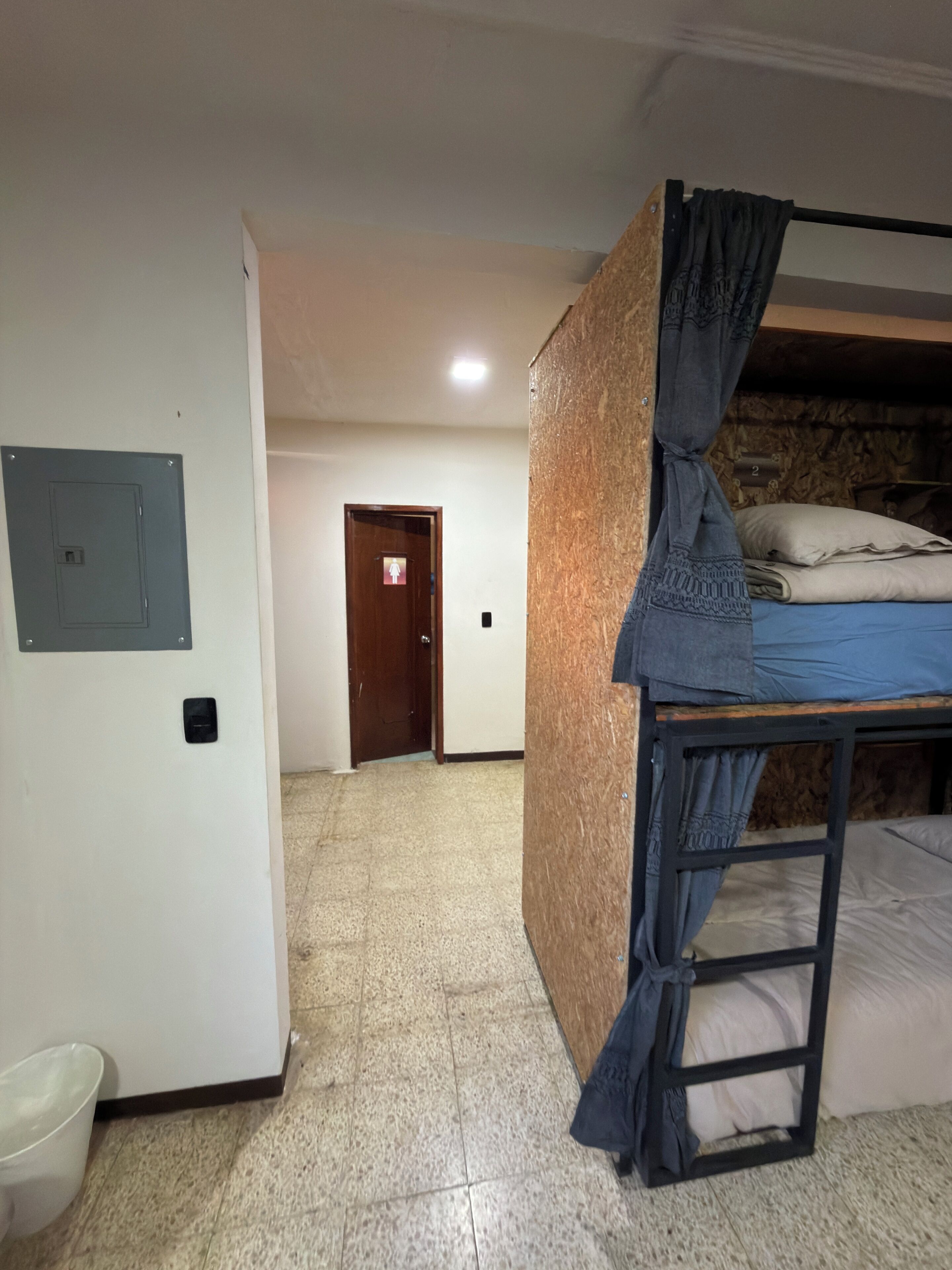 Une pièce avec un lit superposé, une porte avec un panneau indiquant la sortie, un luminaire fixé au mur et une poubelle.