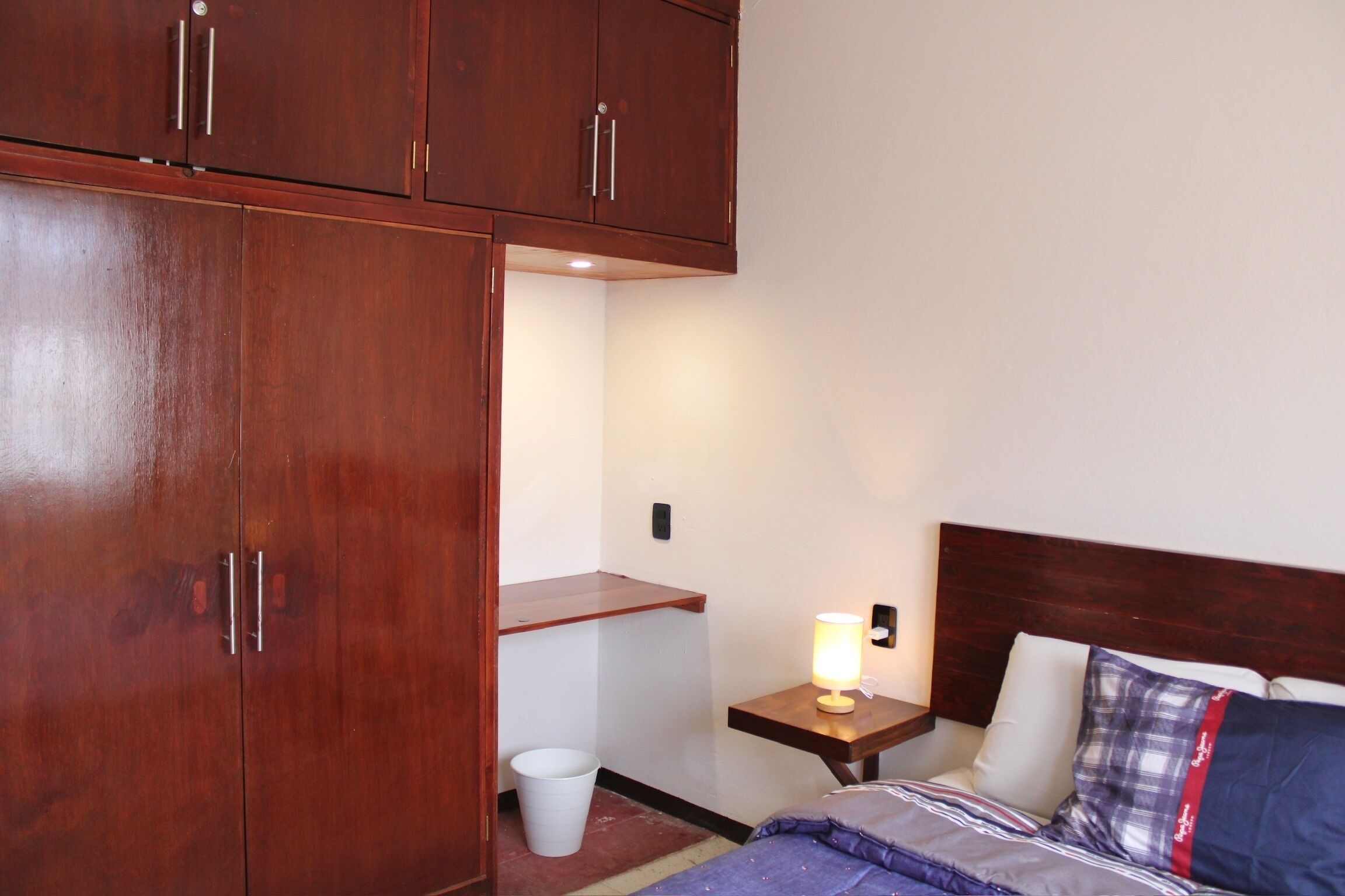 A bedroom with a bed, a nightstand with a lamp, a wooden wardrobe, and a small white trash bin.