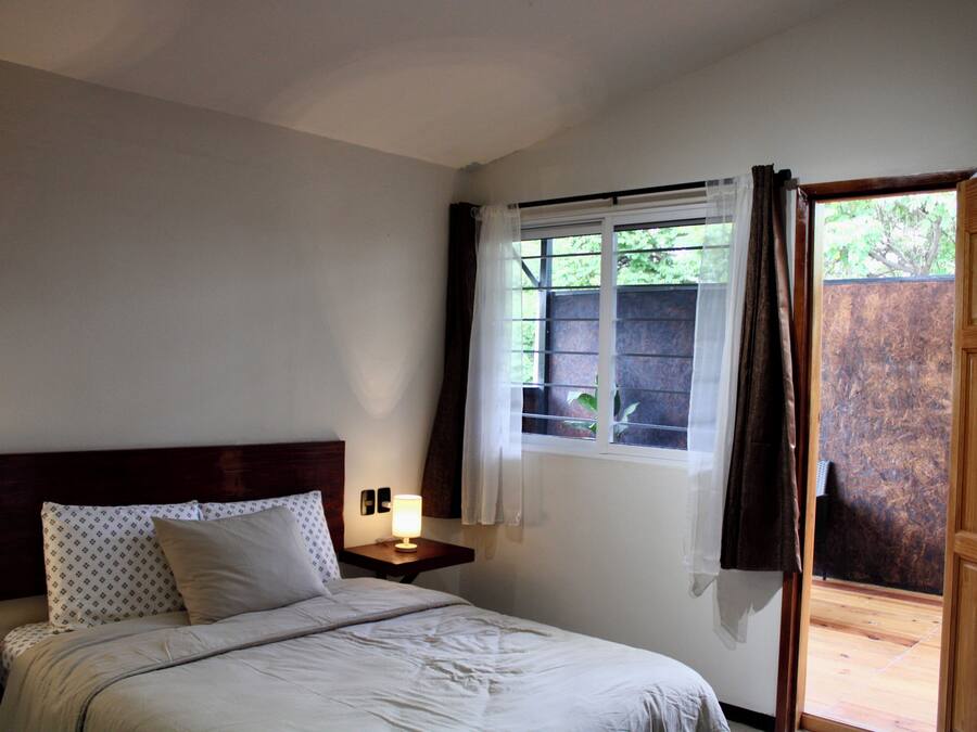 A bedroom with a bed, a bedside table with a lamp, a window with curtains, and a wooden door.