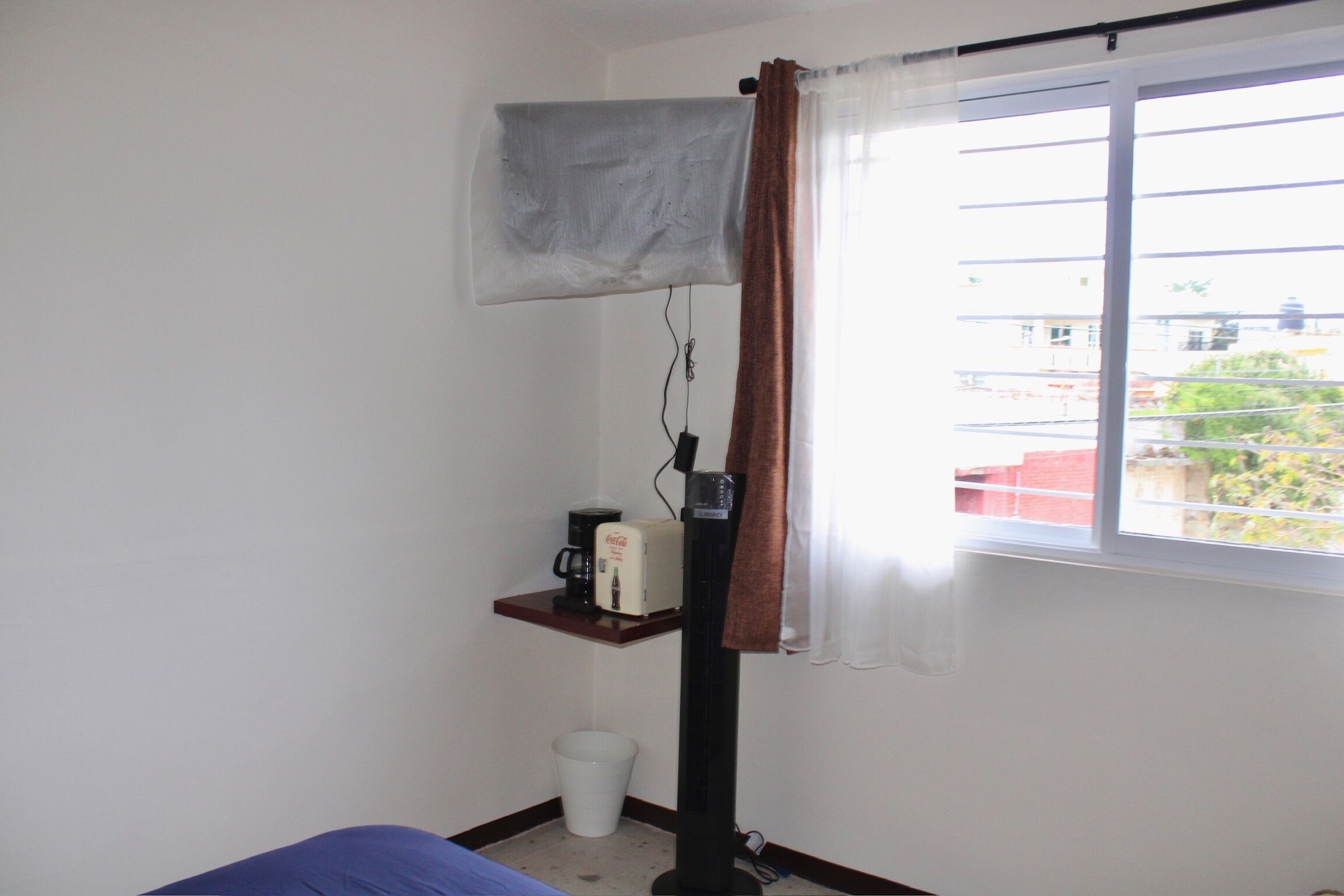 Une chambre avec un lit, un radiateur mobile, une machine à café, une poubelle et une fenêtre donnant sur des immeubles.