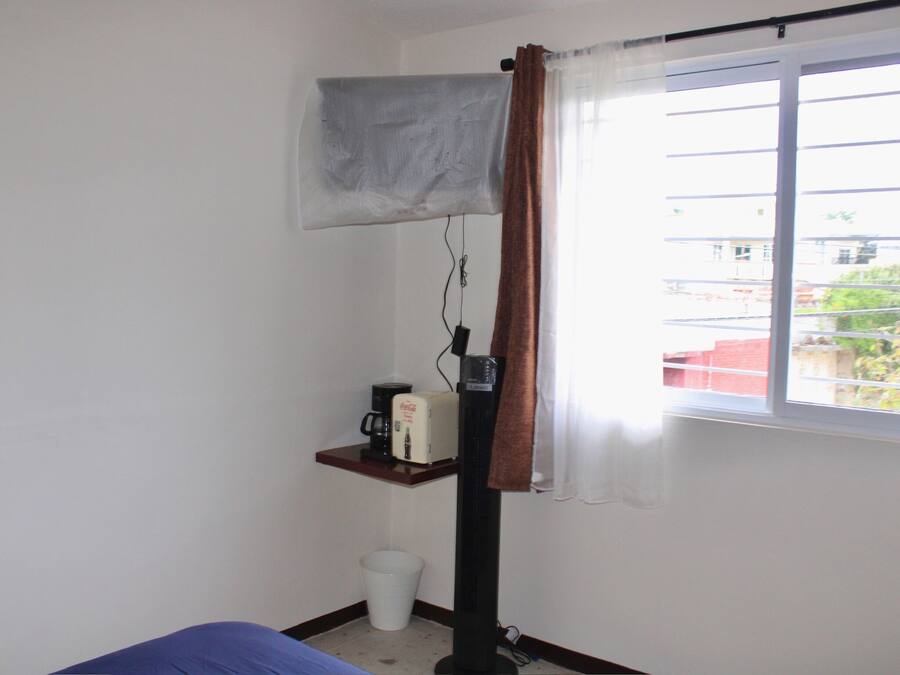 Une chambre avec un lit, un radiateur mobile, une machine à café, une poubelle et une fenêtre donnant sur des immeubles.