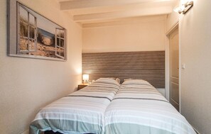 Ein schmales Zimmer mit einem Bett, einem Nachttisch, einer Lampe und einem gerahmten Bild an der Wand.