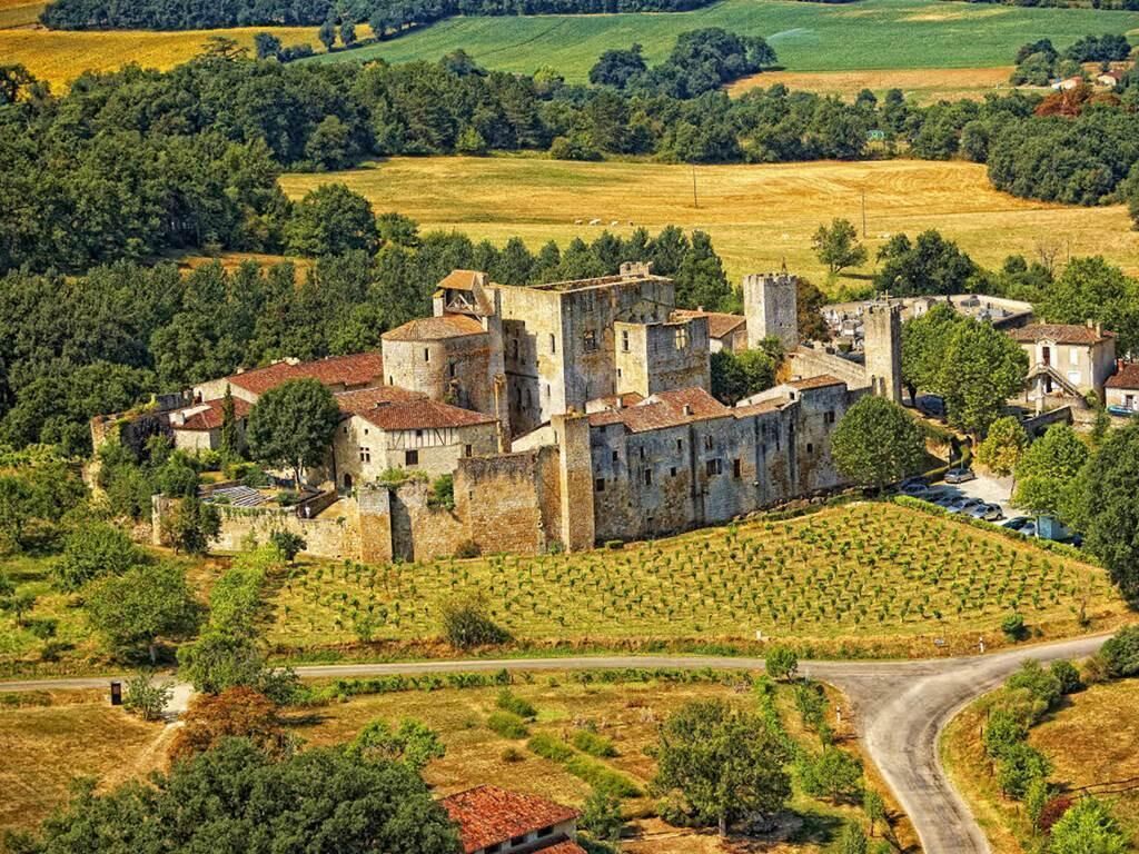 A historic stone castle with multiple towers, surrounded by lush greenery and agricultural fields.