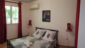 A bedroom with a bed, a window with curtains, an air conditioner, a wall-mounted artwork, and a bedside table with a lamp. - MA Season, 2 detached four-bedroom villas for groups or families of 6 to 12 people (Sainte-Luce)