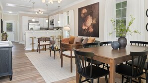 Una cocina moderna con armarios blancos, un comedor con mesa y sillas de madera, y un cuadro grande con motivos florales en la pared.