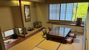A tatami mat room with a wooden table and chairs, a shoji screen, and a view of greenery outside.