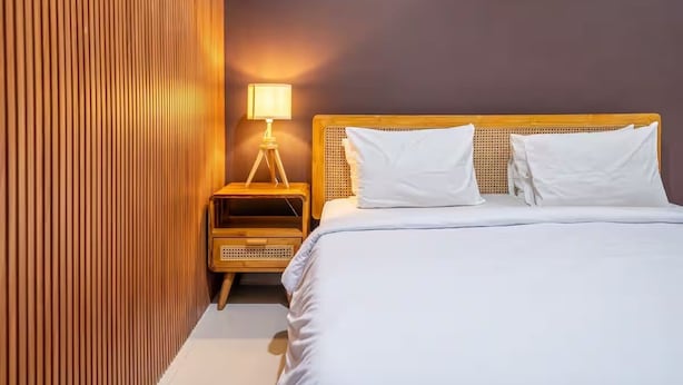 Una cama bien hecha con sábanas blancas, cabecera de madera y mesita de noche con lámpara.