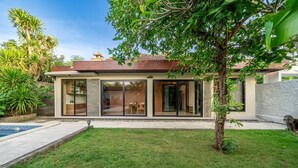 Una casa moderna con piscina, un césped cuidado y un gran árbol en el jardín delantero.