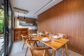 Uma sala de jantar com uma mesa e cadeiras de madeira, uma cozinha ao fundo e uma porta de vidro deslizante que dá para uma área externa.