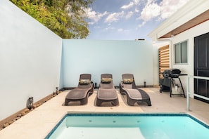 A poolside area with three lounge chairs, a barbecue grill, and a clear blue sky.